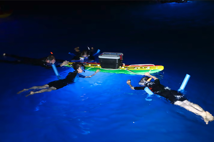Snorkelers float in the ocean at night next to surf board with lights underneath. 
