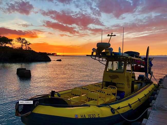 A yellow boat sits next to a dock with a colorful sunset over the ocean behind it. 