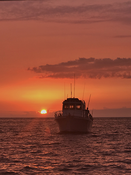 A ship floats in the ocean under an orange ski with sun setting behind it. 