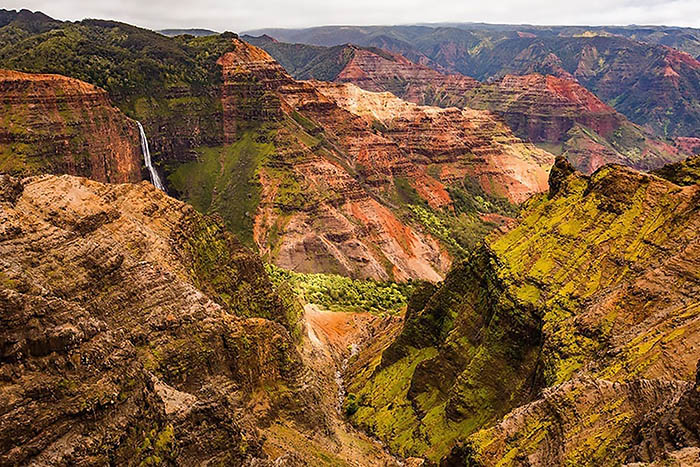 A view of the Waimea Canyon on Kauai, with red cliffs and a waterfall in the distance. 