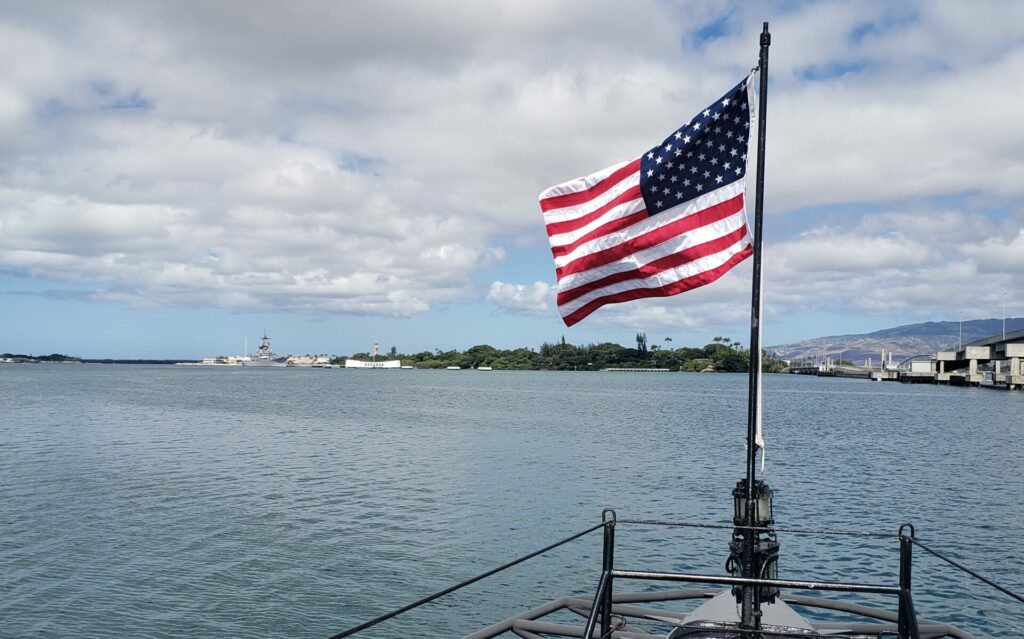 A flag flys from the bow of the USS Bowfin submarine, with the USS Arizona Memorial and the USS Missouri Battleship in the distance, a common sight when you visit Pearl Harbor.